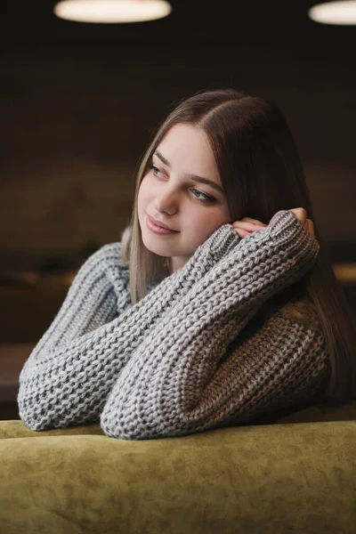 Young female in knitted cardigan of grey color sitting with pensive look and thinking of something