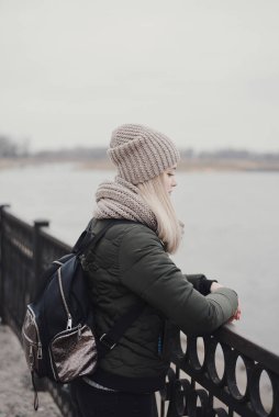 Sarı saçlı, haki rengi ceketli, atkı ören, şapkalı ve sırt çantalı genç bir kadın rıhtımda duruyor ve yağmurlu bir günde nehire bakıyor.