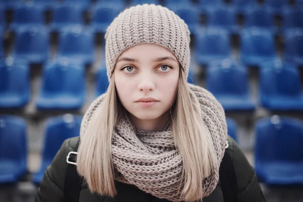 一个金发碧眼的少女站在室外 戴着针织围巾和帽子 背景为蓝色体育场座位的画像 — 图库照片
