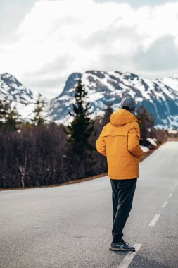 Genç bir adam karlı dağların arka planında yolda duruyor.