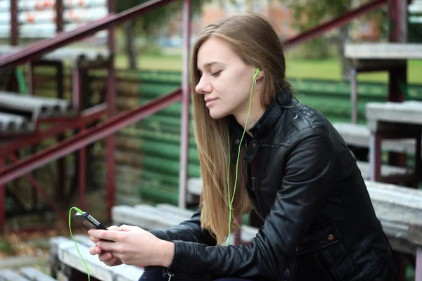 Ung smuk pige ser og lytter musik på din mobiltelefon de gamle stadioner bænk - Stock-foto