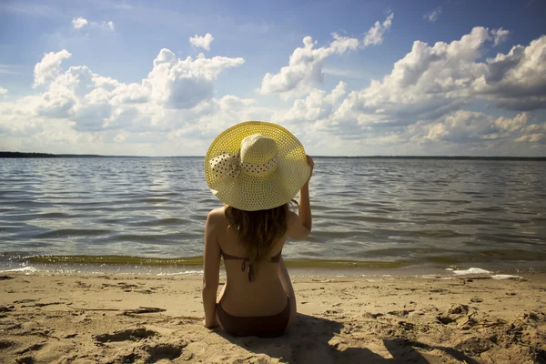 Ung vacker flicka i stråhatt sitter på gul sand nära havet Royaltyfria Stockbilder