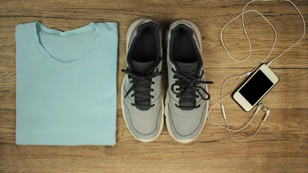 Комплект для спорту: взуття, футболка, мобільний телефон з навушниками крупним планом на дерев'яному фоні, вид зверху — стокове фото