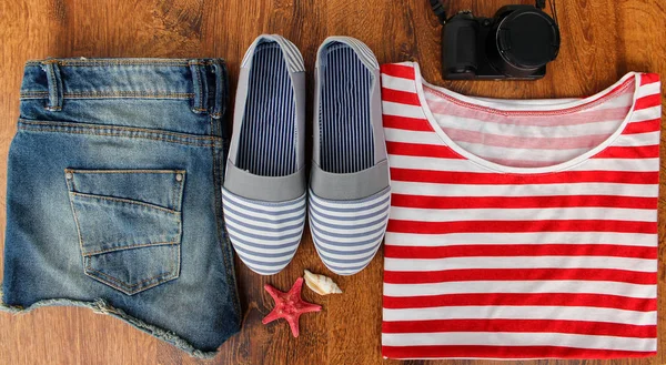 Conjunto de roupas para ir ao mar: calções de ganga, uma camisa listrada e tênis listrados, fotocâmera, conchas, uma visão superior de um fundo de madeira . — Fotografia de Stock