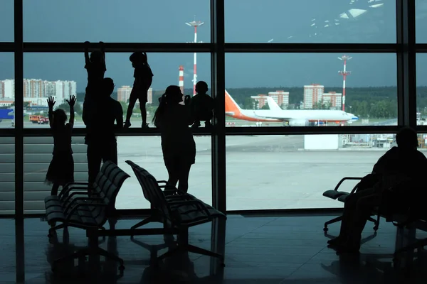 Silhouetten von jungen Familien, die am Fenster stehen und auf die Landebahn des Flughafens mit Flugzeugen blicken und auf ihren Flug warten — Stockfoto