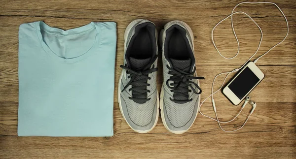 Комплект для спорта: обувь, футболка, мобильный телефон с наушниками крупным планом на деревянном фоне, вид сверху — стоковое фото