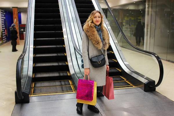 Menina bonita nova é feliz contra um fundo de uma escada rolante em um centro comercial com sacos de compras — Fotografia de Stock