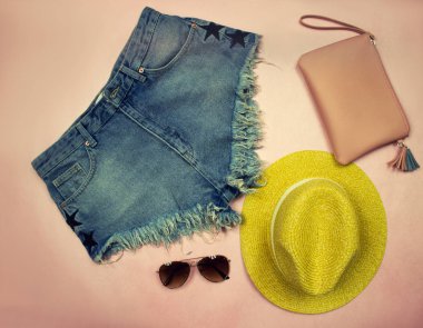 Konu fotoğrafçılık. Giysiler üzerinde kot şort, sarı şapka, gözlük ve kozmetik çantası oluşan bir pembe dizi yaz