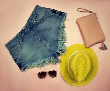 Konu fotoğrafçılık. Giysiler üzerinde kot şort, sarı şapka, gözlük ve kozmetik çantası oluşan bir pembe dizi yaz