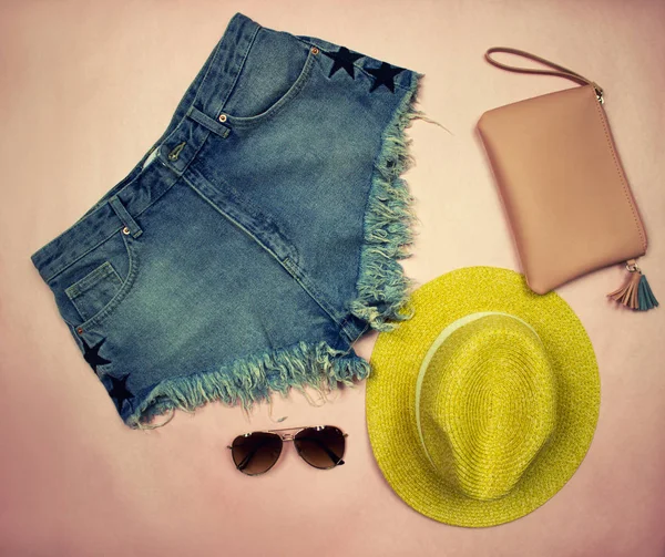 Konu fotoğrafçılık. Giysiler üzerinde kot şort, sarı şapka, gözlük ve kozmetik çantası oluşan bir pembe dizi yaz