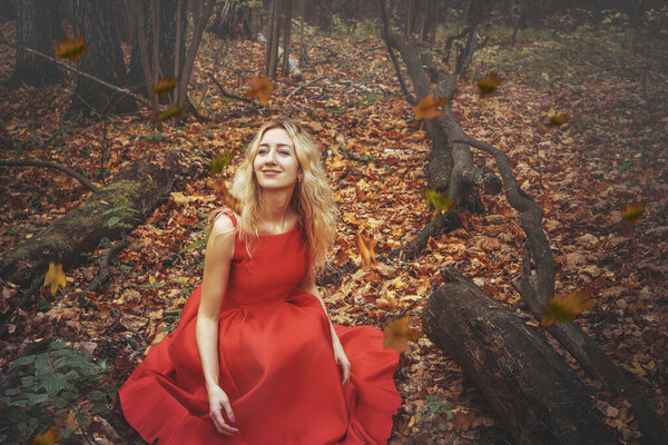 Young pretty woman in the red dress is walking in the foggy mystical forest with fallen leaves.