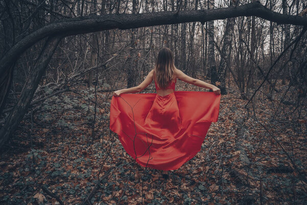 Young beautiful woman in the long red dress is walking along foggy mysterious forest.