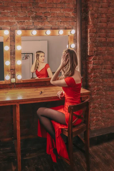 Mladá sexy dívka v červených dlouhých šatech se připravuje na střelbu a dívá se na její odraz v zrcadle šatny — Stock fotografie