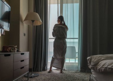 Beyaz bornozlu genç ve güzel bir kadının silueti. Sabahın erken saatlerinde otel odasından deniz manzarasına bakıyor..