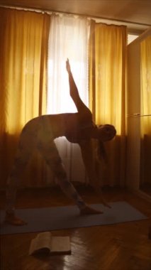 İnce, genç bir kadın sabahın erken saatlerinde arka planda güneş ışığıyla evindeki mavi halıda yoga yapıyor.