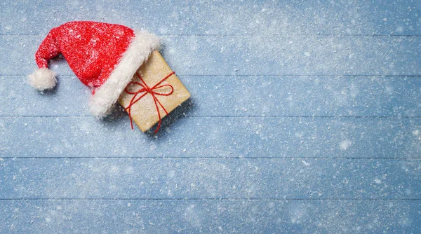 Kahverengi hediye kutusu ahşap masa örtülü kar ile - mavi Noel Baba şapkası ile panorama — Stok fotoğraf