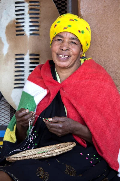 Hermosa mujer zulú africana en ropa étnica está tejiendo una joyería de cuentas. Traje tradicional hecho a mano, tocado y manta . — Foto de Stock