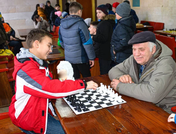 Büyükbabam ve torunum satranç oynuyorlar. Öğrenciler arasında satranç turnuvası. Çocuklar satranç oynar. Karnaval kutlaması. Bilogorotka. Ukrayna. Shrovetide, 25 Şubat 2017
