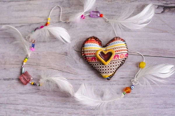 Textile heart of handmade with ethnic ornament on a grayish old aged background. decorative element of white feathers. Ceramic heart as an element of decor. Valentine's Day. — Stock Photo, Image