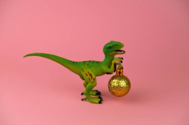 Yumuşak pembe arka planda altın noel topu olan komik plastik dinozor.