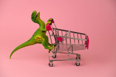 Yumuşak pembe arka planda alışveriş arabası olan komik yeşil dinozor oyuncağı.