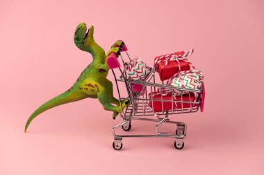 Komik yeşil dinozor oyuncağı. Alışveriş arabası dolusu hediye kutusu ve yumuşak pembe arka planı var.