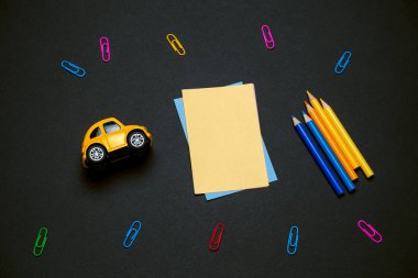 Boş sarı bir kağıt, kalem, ataç ve siyah bir karatahtanın üzerinde sarı bir oyuncak araba, okula dönüş konsepti.