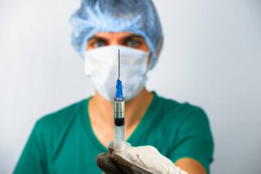 Şapkalı, maskeli ve eldivenli bir erkek doktor elinde şırıngayla arka planda bulanık bir adamın şırıngasıyla elini kapatıyor.