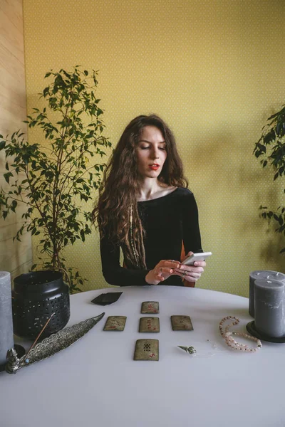 beautiful young woman in a black corduroy dress is guessing on cards with runes and uses an online app in phone to interpret divination