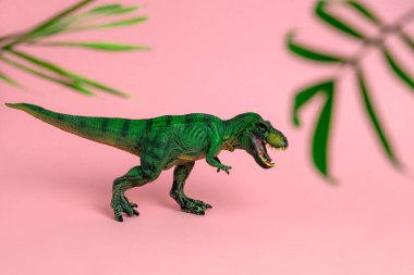 Açık ağızlı yeşil dinozor oyuncağı. Pembe arka planda bulanık palmiye yaprakları arasında.