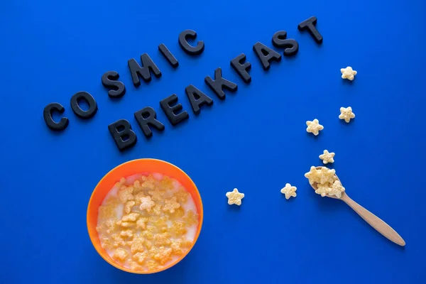 top view flat lay wooden bowl with crunchy corn stars and milk  and inscription cosmic breakfast on a blue background , space day card