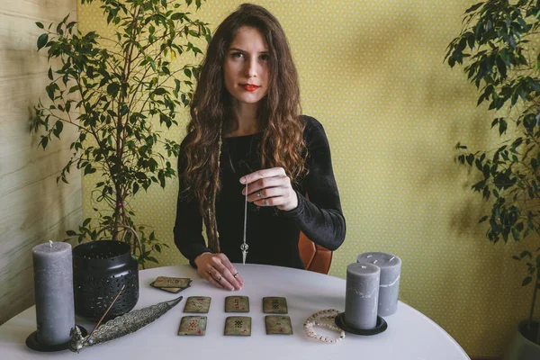 Tyumen, Rusya, 12 Şubat 2020. Siyah kadife elbiseli güzel bir kadın ruhani bir seans sırasında rünlerin üzerinde gümüş bir zincir tutuyor.