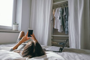 Genç esmer kadın yatakta uzanıyor ve akıllı telefon kullanıyor. Kadın evde akıllı telefon kullanıyor. Yatak odasında.