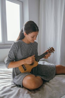 Ukulele oynayan güzel genç bir kadın yatak odasında yatakta oturuyor hobileri ve müzik eğitimi konsepti.