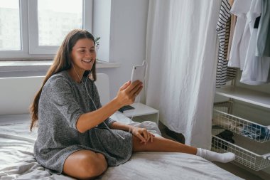 Genç, güzel, pozitif, uzun siyah saçlı, gri elbiseli, kulaklık takan, cep telefonlu bir kadın.