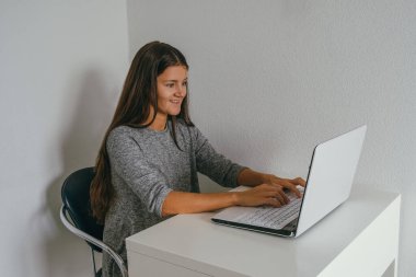 Gündelik gri elbise giyen genç ve güzel bir kadın beyaz duvara karşı dizüstü bilgisayarla çalışıyor.