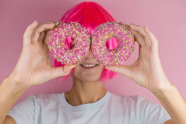 粉红头发的年轻妇女 粉红背景上有两个粉红甜甜圈 用女性的手把釉面甜甜圈关上 — 图库照片