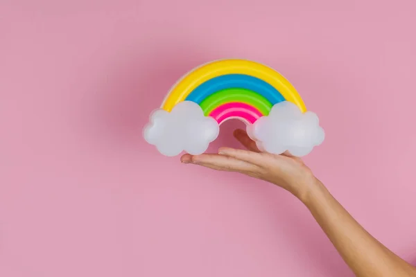 用粉红底色的云彩握住塑料彩虹的女性手 — 图库照片