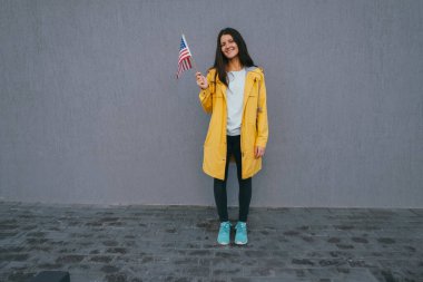 Mutlu, güzel, esmer bir kadın gülümsüyor ve gri duvarda Amerikan bayrağı taşıyor.