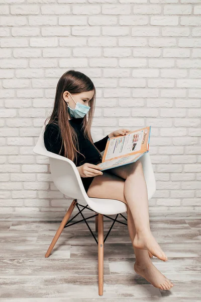 The girl in a blue mask is in quarantine at home.The girl sits on a modern white chair and reads a book. White brick wall. Isolation of the house on the coronavirus Spread and pandemic in the world. Somber woman in shelter on Curfews time around city