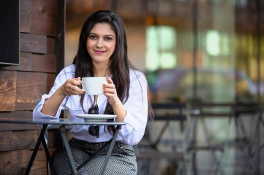 Güzel esmer, Amerikalı kadın kahve dükkanının avlusunda cappuccino yudumluyor, kahve fincanıyla rahatça gülümsüyor.