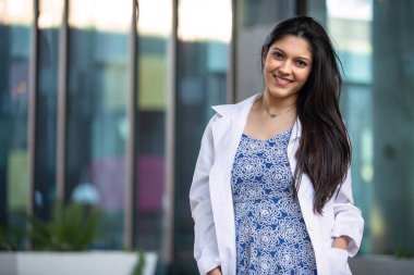 Özel muayenehanenin önünde duran beyaz önlüklü çok ırklı kadın doktor, muhtemelen doktor, diş hijyenisti, terapist, psikiyatrist.
