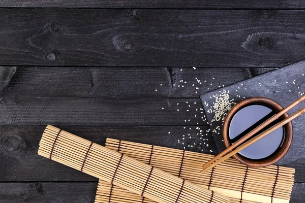 Bakgrunden för sushi. Bambu matta, soja, pinnar på mörka bord. Ovanifrån med kopia utrymme — Stockfoto