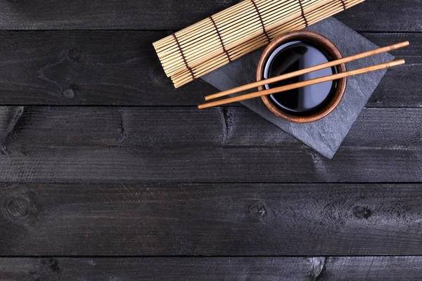 Bakgrunden för sushi. Bambu matta, soja, pinnar på mörka bord. Ovanifrån med kopia utrymme — Stockfoto