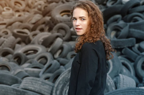 Krásná dívka, kudrnaté vlasy, portrét na pozadí skládky staré auto pneumatiky. Dívá se doleva. — Stock fotografie