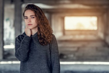 Kıvırcık saçlı. Terk edilmiş bir beton binanın arka planında güzel bir kız. Endüstriyel bölgede arkadan gelen ışık.