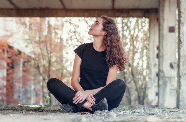 Uzun kıvırcık saçlı hoş bir kız eski bir beton binada bacak bacak üstüne atmış oturuyor. Sola bak..