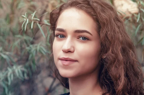 Portrait of girl with brown curly hair on background of green bush sea buckthorn, close-up face — Stock Photo, Image