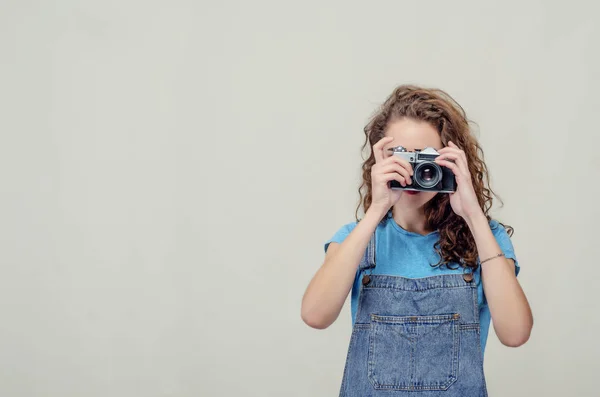 Ragazza bruna riccia in tuta di jeans sta tenendo una macchina fotografica vintage tra le mani. Scatta una foto — Foto Stock