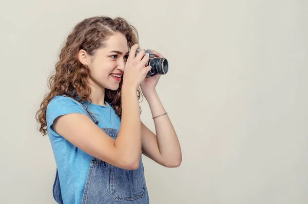 Ragazza bruna riccia in tuta di jeans sta tenendo una macchina fotografica vintage tra le mani. Scatta una foto. Vista laterale . — Foto Stock